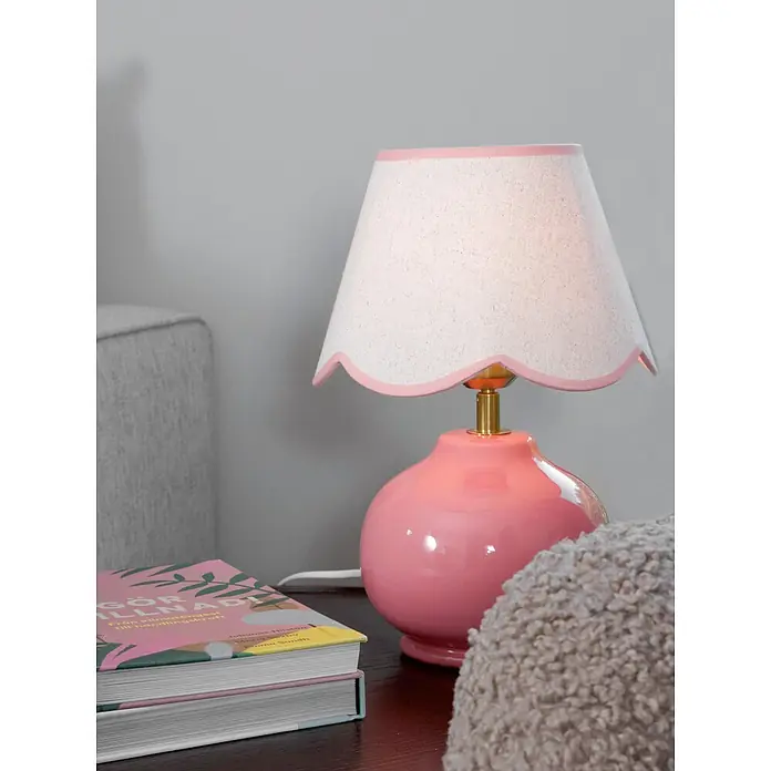 PR Home Tischleuchte Nell - 40 W, E27 - Glänzende rosa Tischlampe mit Messingdetails und einem hellen Stoffschirm mit Wellenkante, die eine Nachttischumgebung beleuchtet.