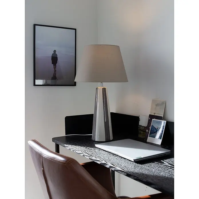 PR Home Tischleuchte Leya - 25 W, E27 - Eine moderne Schreibtischlampe mit hellgrauem Stoffschirm und gestreiftem Keramikfuß, die einen Eckschreibtisch mit Laptop beleuchtet.