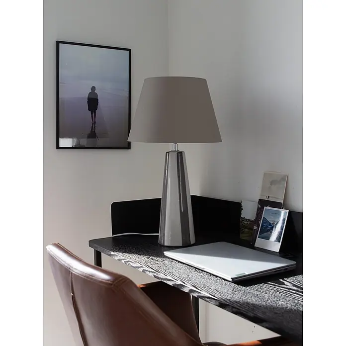 PR Home Tischleuchte Leya - 25 W, E27 - Eine moderne graue Tischlampe mit gestreiftem Keramikfuß und grauem Schirm steht auf einem dunklen Schreibtisch neben einem geschlossenen Laptop und einem braunen Ledersessel.