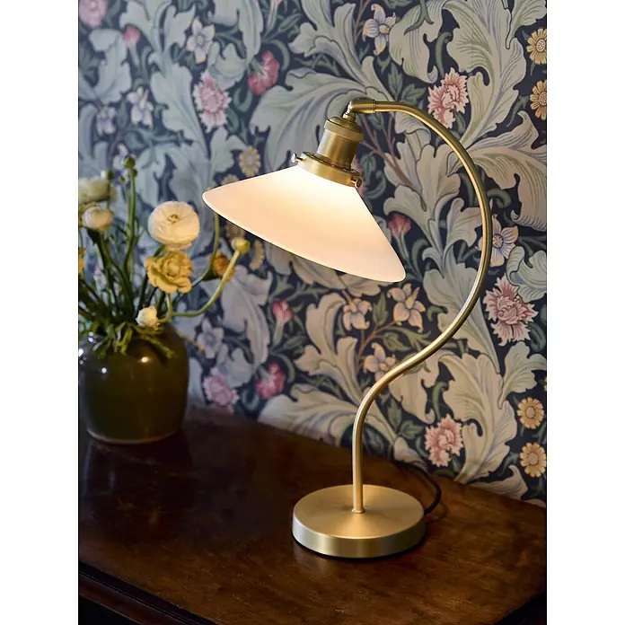PR Home Tischleuchte Axel - 15 W, E27 - Messing-Tischlampe mit mattem Glasschirm, beleuchtet auf dunklem Holztisch vor Blumentapete.