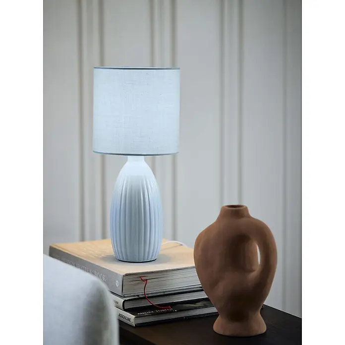PR Home Tischleuchte Uno - 25 W, E14 - Weiße gerippte Keramik-Tischlampe mit hellblauem Schirm auf Büchern neben einer braunen Vase.