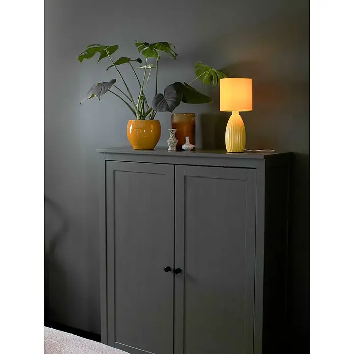 PR Home Tischleuchte Uno - 60 W, E14 - Ein dunkelgrauer Schrank dient als Ablagefläche, darauf stehen eine Pflanze, eine Lampe und Dekoartikel in einem Wohnraum.