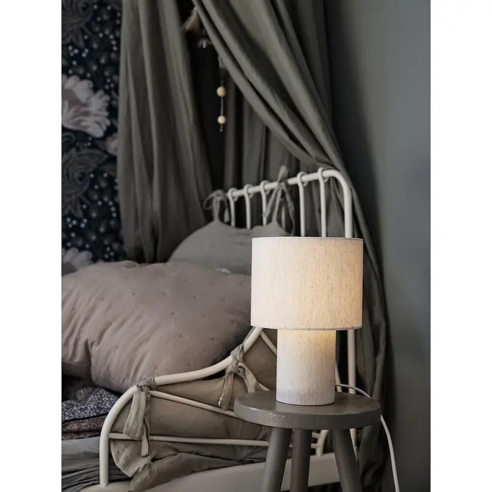PR Home Tischleuchte Leah - 15 W, E27 - Weiße Stofftischlampe mit hellem Sockel auf grauem Hocker, beleuchtet einen Nachttischbereich.
