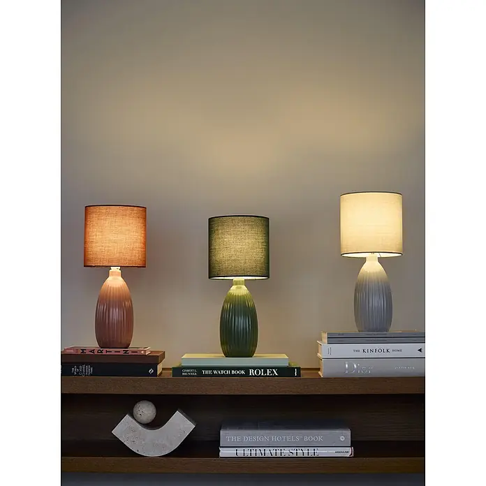 PR Home Tischleuchte Uno - 25 W, E14 - Drei moderne Keramiktischlampen mit Textilschirmen in Terrakotta, Grün und Hellgrau, auf einem Holzregal mit Büchern.