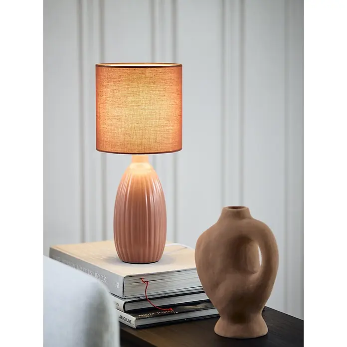 PR Home Tischleuchte Uno - 25 W, E14 - Beleuchtete Terrakotta-Tischlampe mit geripptem Keramikfuß und Stoffschirm, auf einem Stapel Bücher.