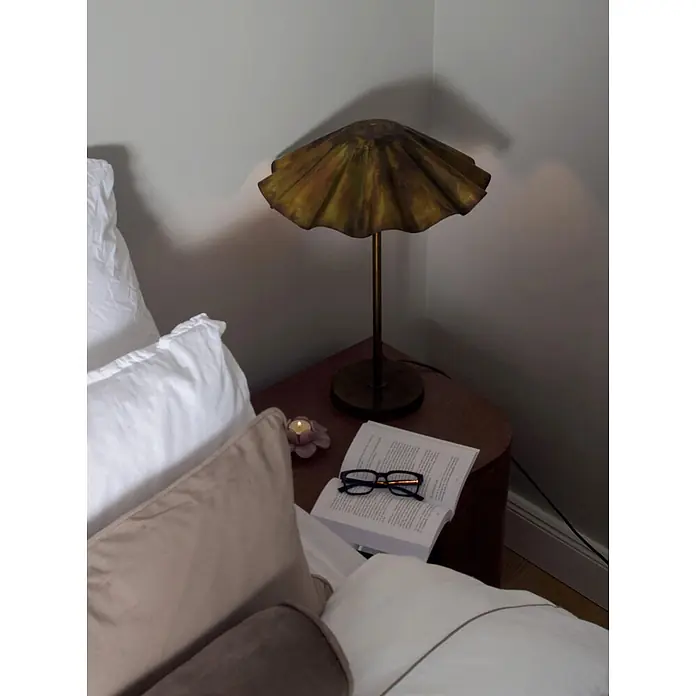 PR Home Tischleuchte Aira - 25 W, E27 - Messingfarbene Rüschenlampe auf dunklem Nachttisch mit offenem Buch, Lesebrille und brennender Kerze am Bett.