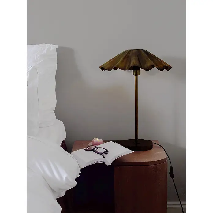 PR Home Tischleuchte Aira - 25 W, E27 - Goldfarbene Tischlampe mit gerafftem Metallschirm auf einem Nachttisch neben einem Buch und einer Brille.