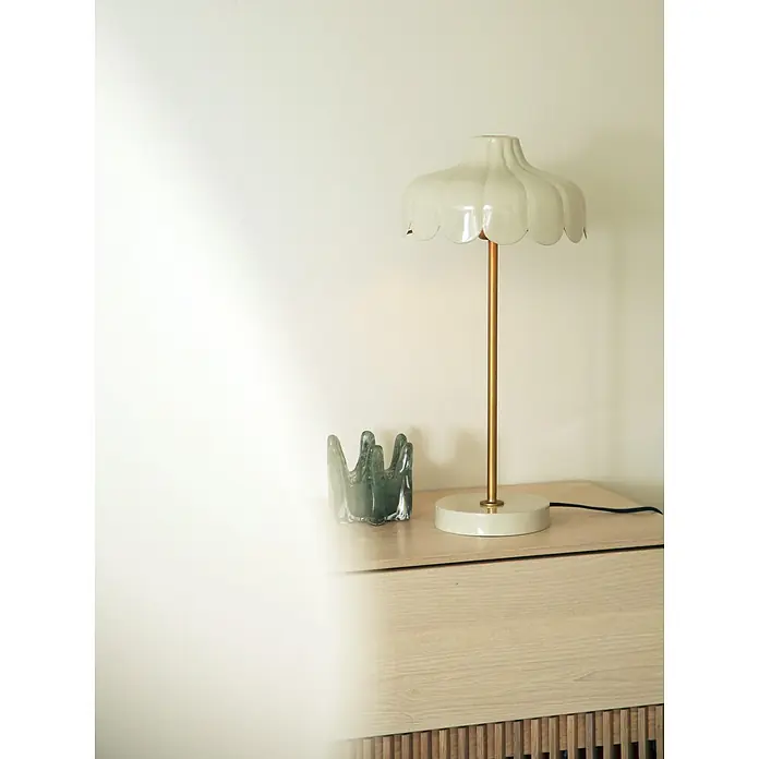 PR Home Tischleuchte Tischleuchte Wells - 25 W, E27 - Eine cremefarbene Tischlampe mit gewelltem Schirm und goldenem Ständer auf einem hellen Holzschrank, neben einer dunkelgrünen gewellten Glasskulptur.