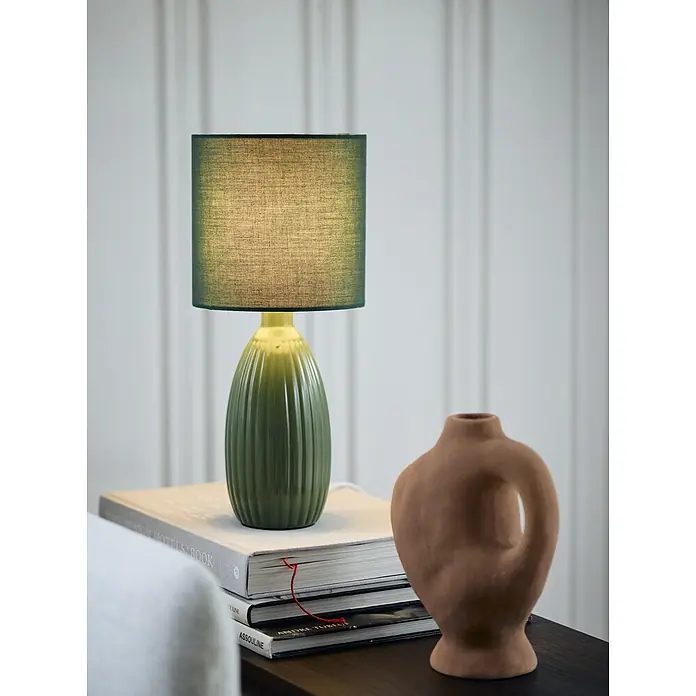 PR Home Tischleuchte Uno - 25 W, E14 - Eine grüne Tischlampe mit geripptem Keramikfuß und strukturiertem Stoffschirm, auf Büchern neben einer Terrakottavase.