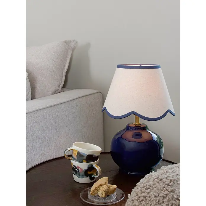 PR Home Tischleuchte Nell - 40 W, E27 - Blaue Keramik-Tischlampe mit gewelltem cremefarbenem Leinenschirm auf dunklem Beistelltisch neben hellgrauem Sofa. Gestapelte Tassen und Biscotti in der Nähe.