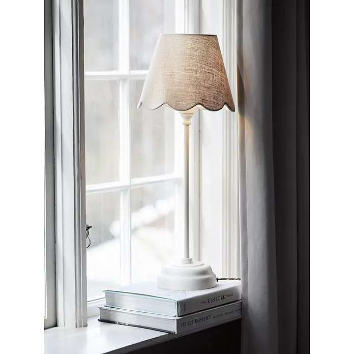 PR Home Tischleuchte Lottie - 40 W, E27 - Weiße Tischlampe mit einem gewellten Leinenlampenschirm, auf Büchern auf einer Fensterbank.