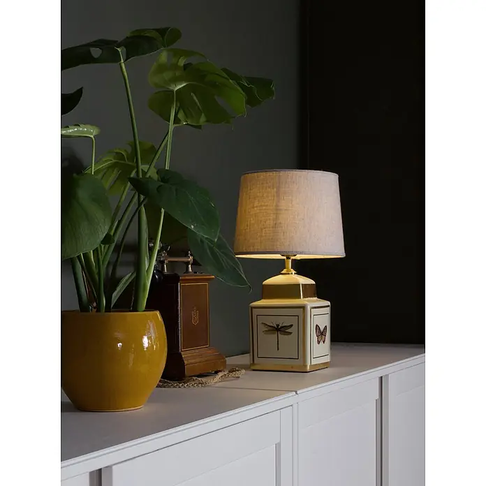 PR Home Tischleuchte Lucca - 40 W, E27 - Beleuchtete cremefarbene Tischlampe mit Goldakzenten, Insektenillustrationen und hellem Stoffschirm auf einem weißen Schrank.