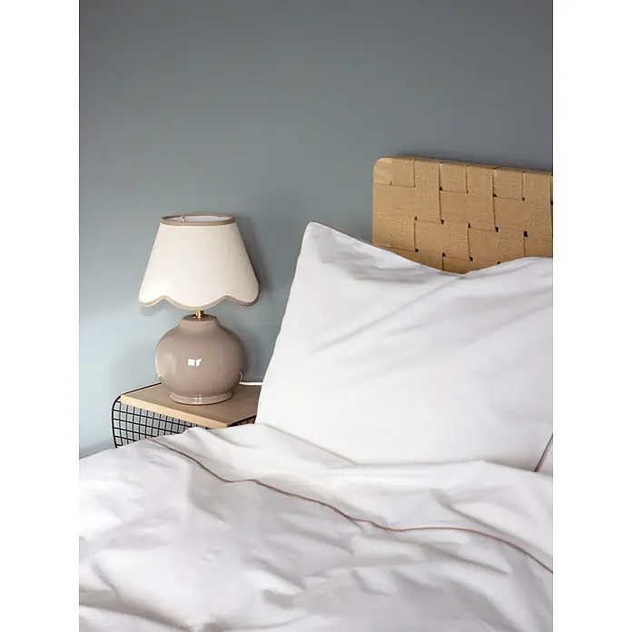 PR Home Tischleuchte Nell - 25 W, E27 - Gemütliches Schlafzimmer mit weißer Bettwäsche, geflochtenem Kopfteil und beigefarbener Lampe mit gewelltem Schirm auf Nachttisch.