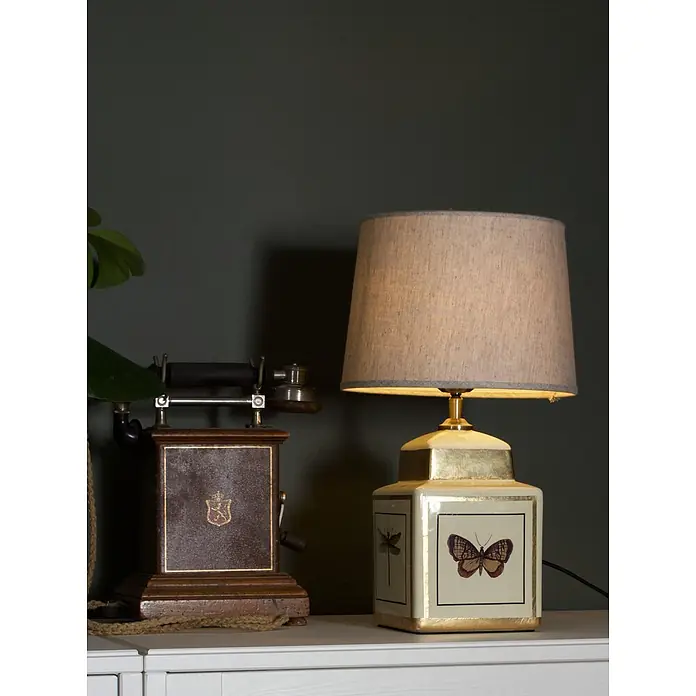PR Home Tischleuchte Lucca - 40 W, E27 - Ein Vintage-Telefon mit braunem Holzsockel und eine Lampe mit beigem Stoffschirm und weiß-goldenem, schmetterlingsverziertem Sockel auf einer weißen Oberfläche.