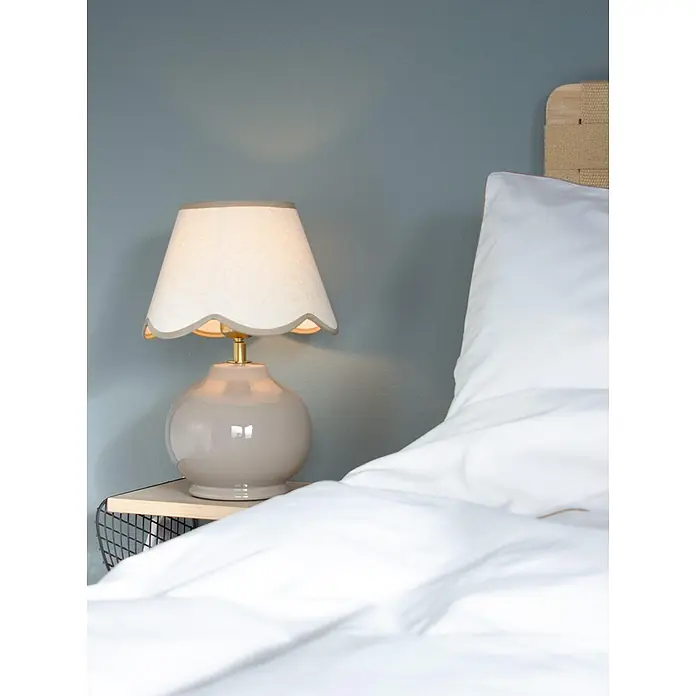 PR Home Tischleuchte Nell - 25 W, E27 - Gezackte beige Nachttischlampe mit glänzendem taupefarbenem Fuß auf Holznachttisch neben weißer Bettwäsche.