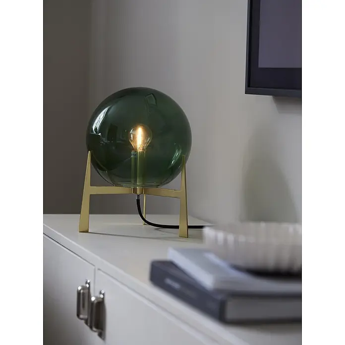 PR Home Tischleuchte Milla - 25 W, E14 - Dunkelgrüne kugelförmige Glastischlampe mit Messingsockel und sichtbarer Glühlampe auf einer Kommode.