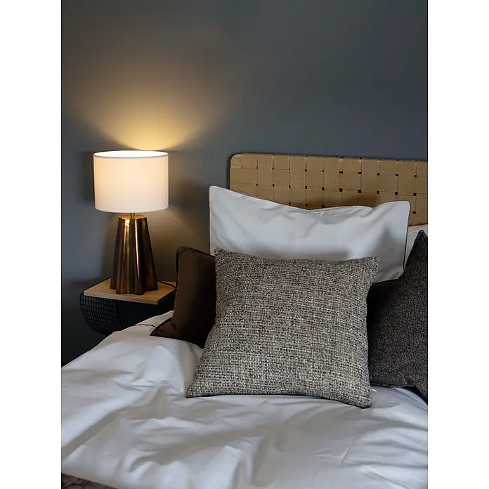 PR Home Tischleuchte 50cm - 40 W, E27 - Bett mit weißer Bettwäsche, braunen und strukturierten grauen Kissen sowie einer Messinglampe auf einem Holznachttisch.