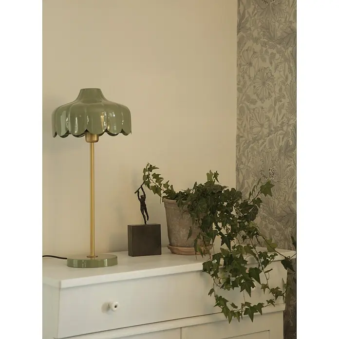 PR Home Tischleuchte Wells - 25 W, E27 - Eine grüne Tischlampe mit gewelltem Schirm und goldenem Stiel steht auf einer weißen Kommode neben einer Blattpflanze.