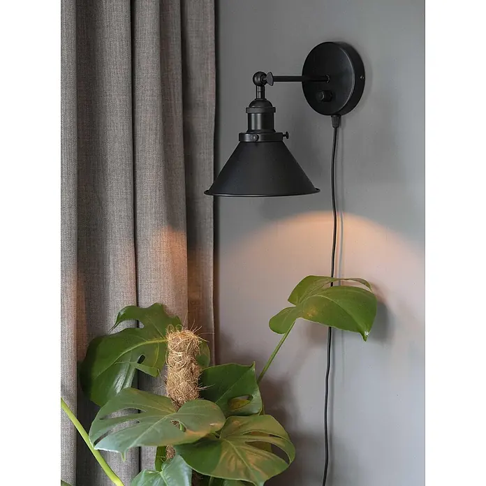 PR Home Wandleuchte Anton - 25 W, E27 - Schwarze Wandlampe aus Metall mit konischem Schirm, beleuchtet eine Monstera-Pflanze. Neben einem grauen Vorhang montiert.