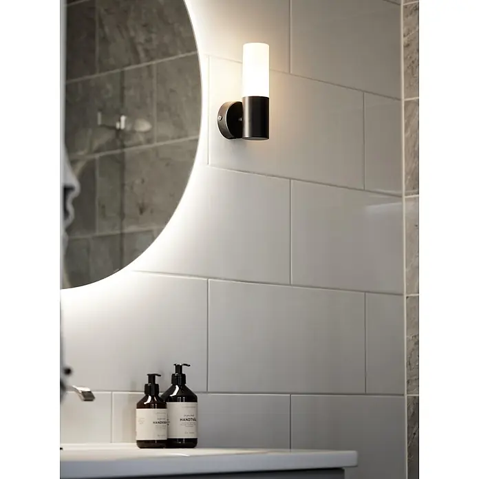 PR Home Wandleuchte Beta - 25 W, E14 - Schwarze Metall-Wandleuchte mit weißem Zylinderschirm, beleuchtet, neben einem beleuchteten Spiegel montiert.