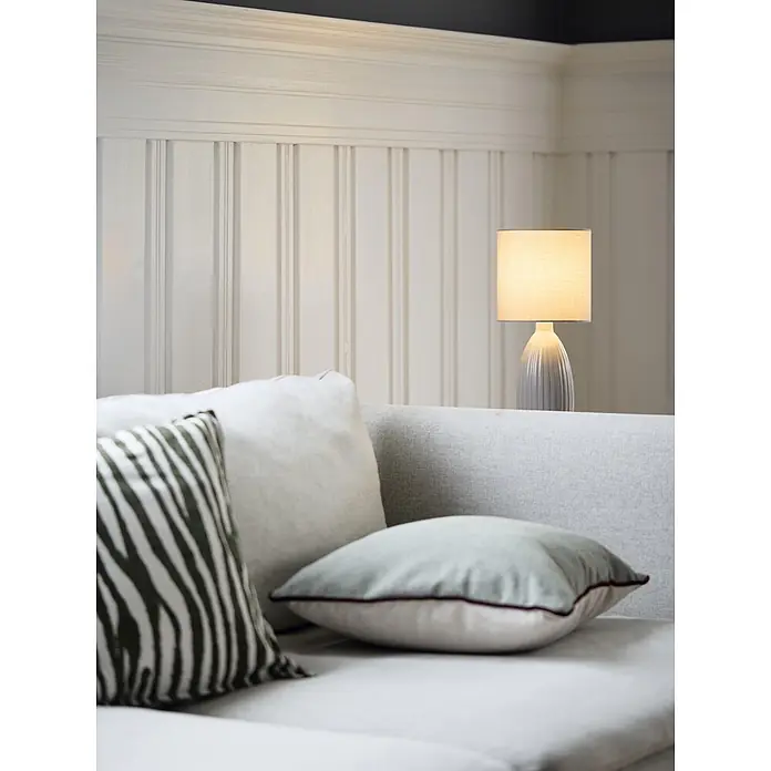 PR Home Tischleuchte Uno - 25 W, E14 - Ein modernes graues Sofa mit Zierkissen, darunter ein grüner Zebramuster, und einer kleinen beleuchteten Tischlampe auf der Rückenlehne.