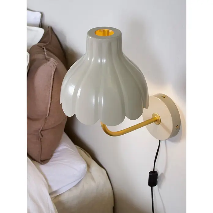 PR Home Wandleuchte Wells - 25 W, E27 - Hellgraue Wandleuchte mit geriffelter Schale und goldenem Arm, an einer cremefarbenen Basis neben einem Bett.