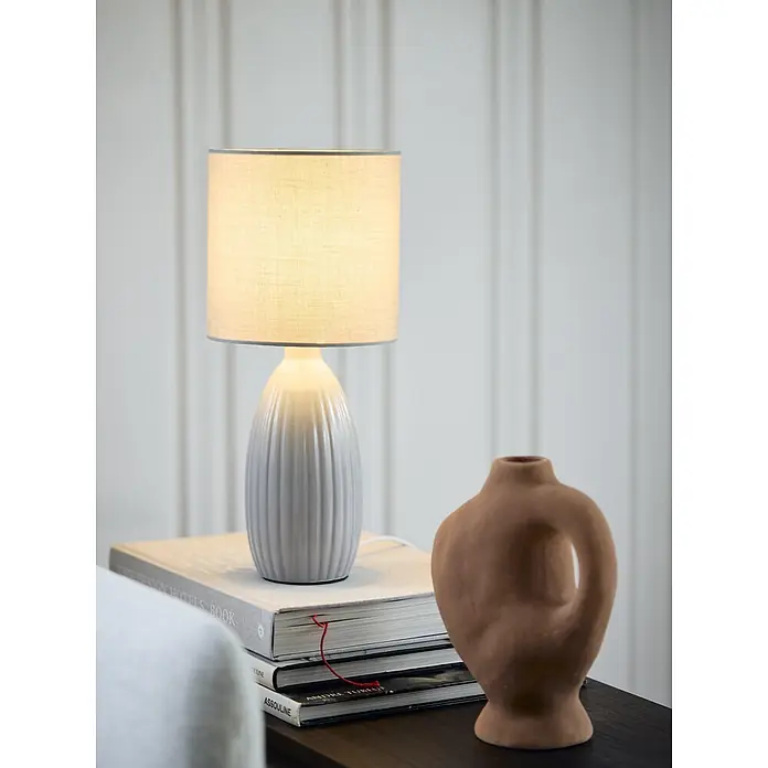 PR Home Tischleuchte Uno - 25 W, E14 - Eine hellgraue Keramik-Tischlampe mit beigem Stoffschirm beleuchtet einen Stapel Bücher auf einem dunklen Tisch, neben einer Terrakotta-Vase.