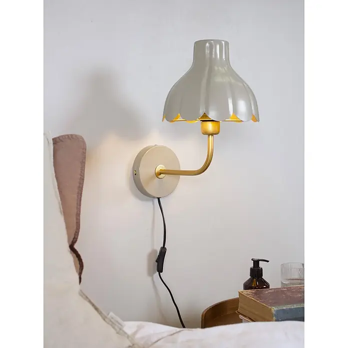 PR Home Wandleuchte Wells - 25 W, E27 - Hellgraue Wandleuchte mit geriffeltem Schirm und Messingarm, über einem Nachttisch montiert. Steckbares Design mit Schnurschalter.