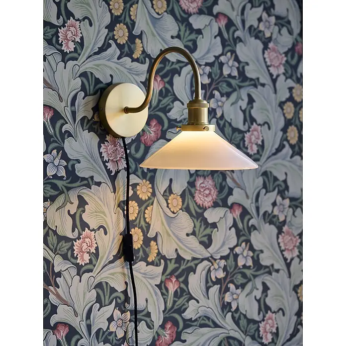 PR Home Wandleuchte Axel - 15 W, E27 - Wandleuchte aus Messing mit weißem, konischem Glasschirm, die warmes Licht vor einer dunklen, gemusterten Tapete abgibt.