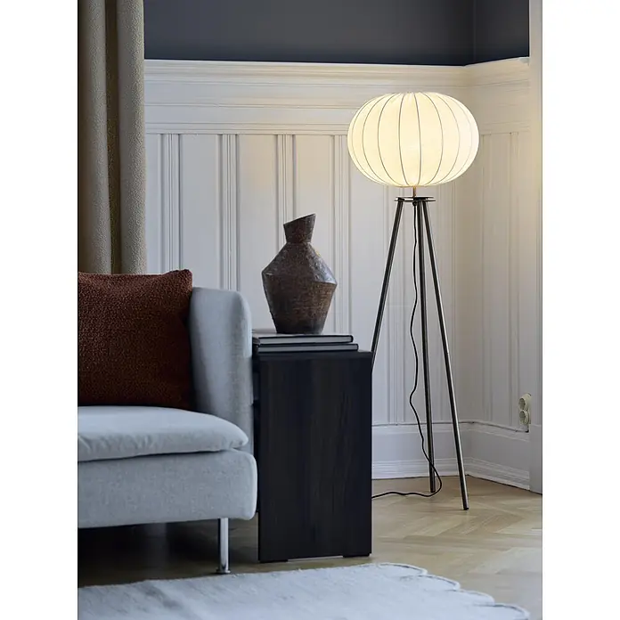 PR Home Stehleuchte Messing - 40 W, E27 - Eine kugelförmige Stoff-Stehlampe mit dunklen Dreibeinständern beleuchtet einen gemütlichen Wohnraum neben einem Sofa.