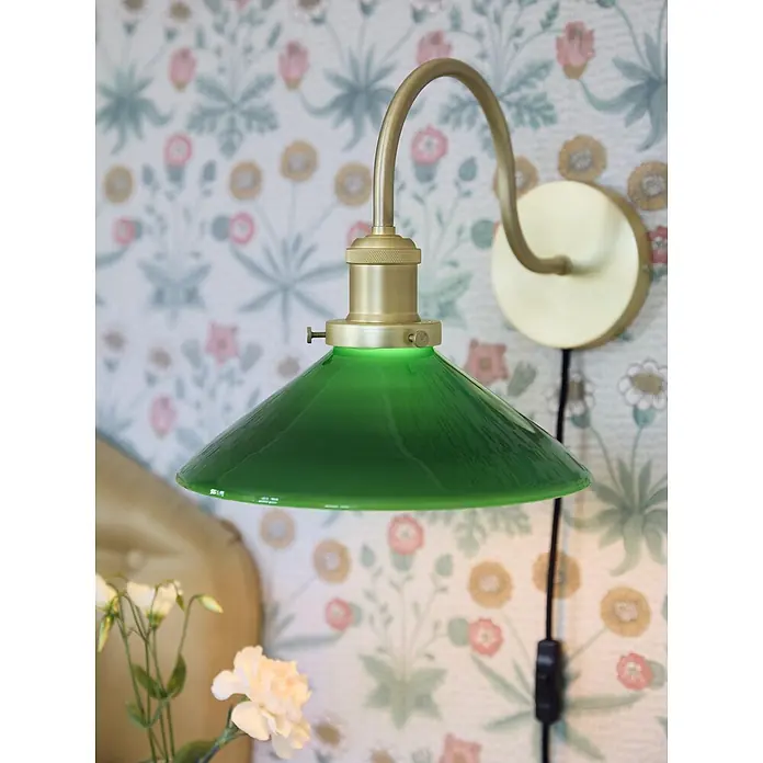 PR Home Wandleuchte Axel - 25 W, E27 - Grüne Glas- und Messing-Wandleuchte mit gebogenem Arm, an einer geblümten Wand montiert.
