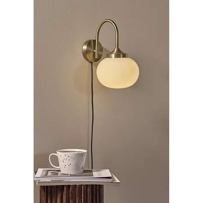 PR Home Wandleuchte Elsy - 240 W, E14 - Wandleuchte aus Messing mit Opalglaskugel, strahlt warmes Licht über einem Nachttisch mit gemusterter Tasse aus.