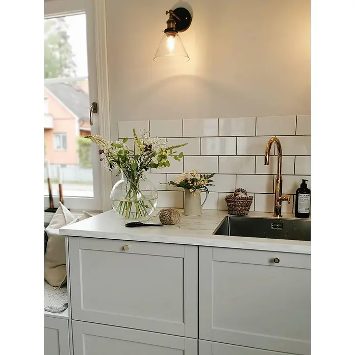 PR Home Wandleuchte Lambda - 15 W, E27 - Helle graue Küchenschränke mit heller Arbeitsplatte, goldener Armatur und Metrofliesen-Spritzschutz um ein Spülbecken.