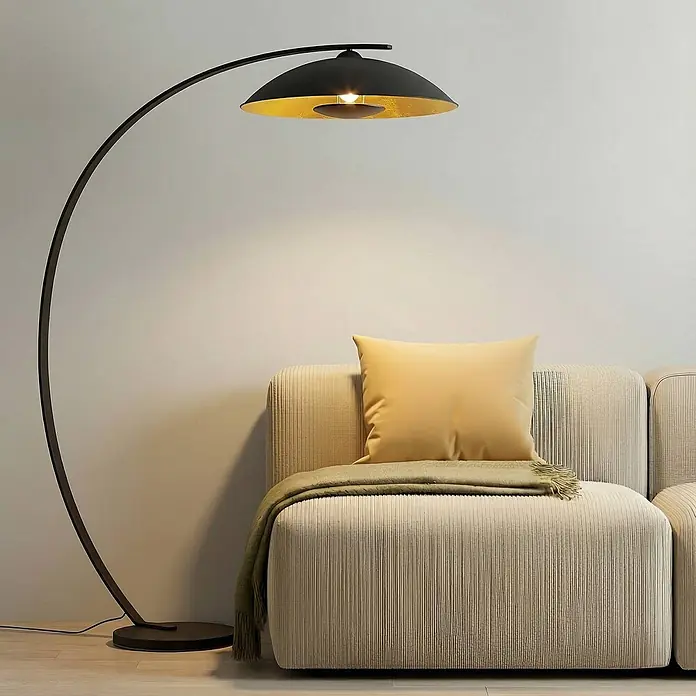 Lindby Stehleuchte Emilienne - schwarz, schwarz, E27 - Schwarze Bogen-Stehleuchte mit goldener Innenschirm, die einen Wohnbereich neben einem beigen Sofa beleuchtet.