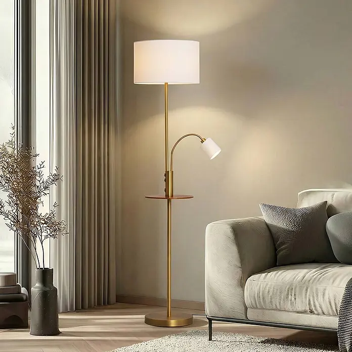 Lindby Stehleuchte Aovan - Höhe: 170 cm, bronze / altmessing, creme / amber, E27 - Goldene Stehlampe mit weißem Schirm, verstellbarer Leselampe und integriertem Holztisch, neben einem Sofa platziert.