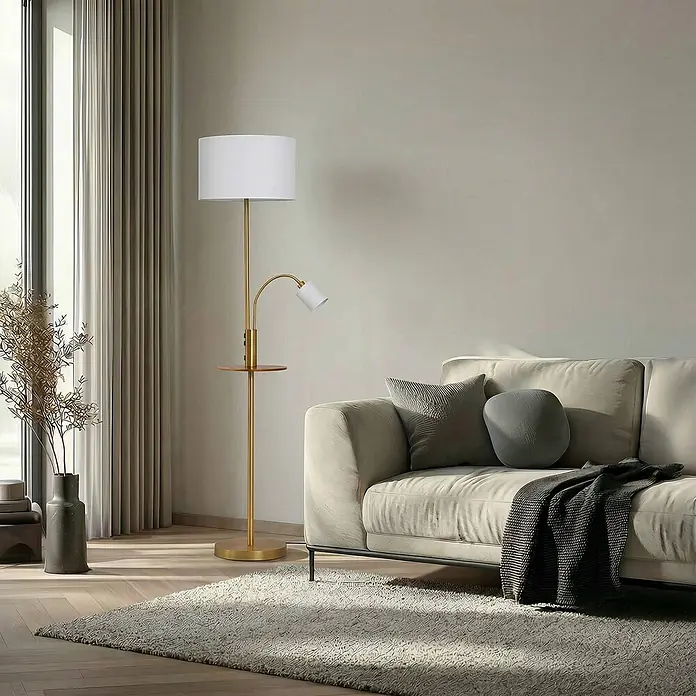 Lindby Stehleuchte Aovan - Höhe: 170 cm, bronze / altmessing, creme / amber, E27 - Goldene Stehlampe mit weißem Schirm, integriertem Holzregal und flexibler Leselampe, neben einem Sofa platziert.
