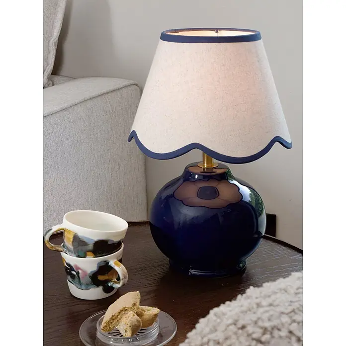 PR Home Tischleuchte Nell - 40 W, E27 - Blaue Keramiktischlampe mit cremefarbenem Wellenrand-Lampenschirm und blauer Bordüre, beleuchtet einen Beistelltisch.