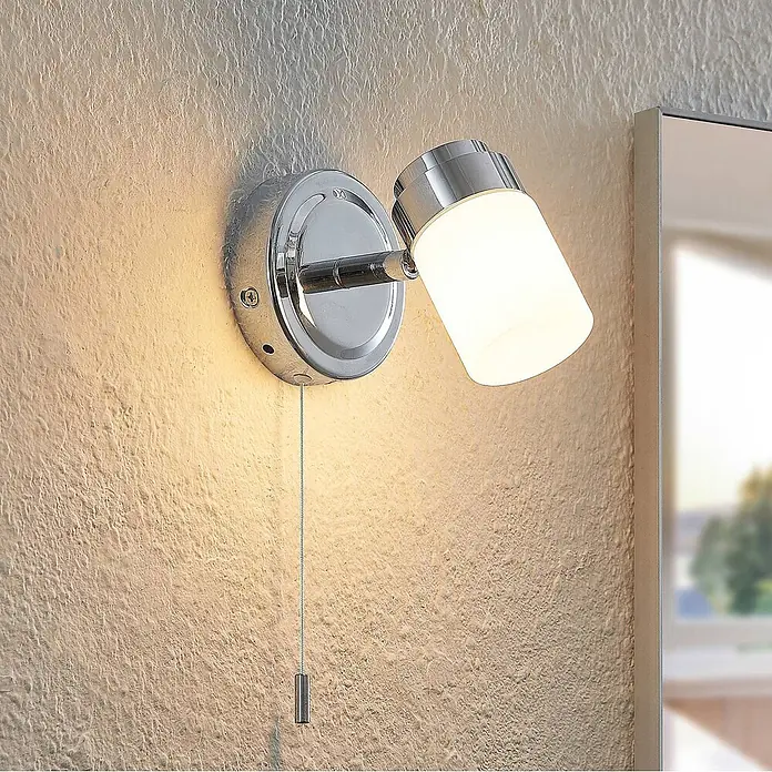 Lindby Wandleuchte Alimari - L x B x H: 12,3 x 9 x 10,6 cm, chrom / silber, G9 - Chrom-Wandleuchte mit weißem zylindrischem Schirm und Zugschalter an einer strukturierten Wand, die den Bereich beleuchtet.
