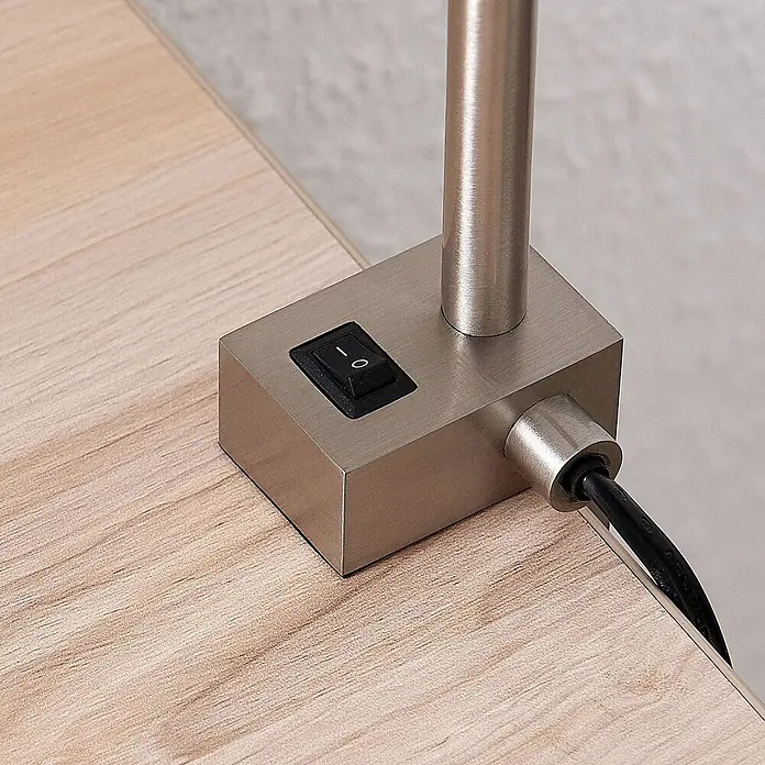 Lindby Klemmleuchte Hanilo - L x B x H: 37 x 4 x 80 cm, Farbe Gehäuse: alu / grau / zink - Lampensockel aus gebürstetem Metall mit schwarzem Ein-/Ausschalter und Netzkabel auf heller Holzoberfläche.