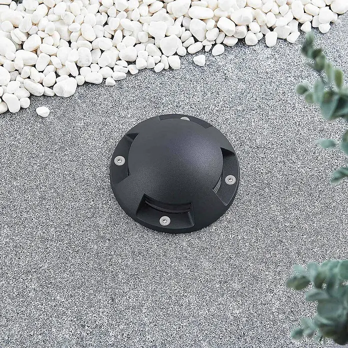 Lindby LED-Gartenstrahler Huban - 3.000 K - Runde schwarze Bodeneinbauleuchte in gesprenkeltem grauem Weg, umgeben von weißen Kieselsteinen und Grün.