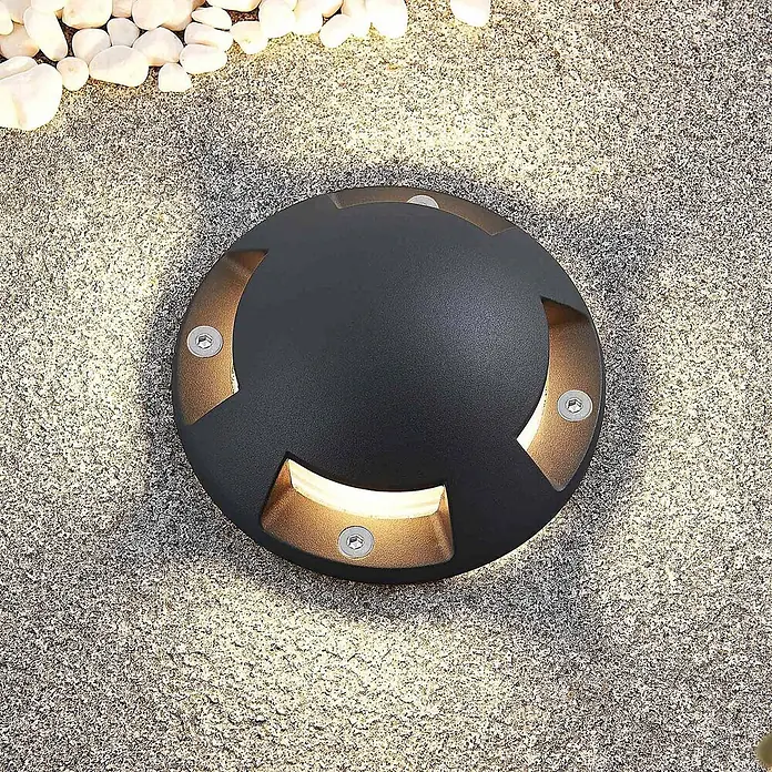 Lindby LED-Gartenstrahler Huban - 3.000 K - Dunkle runde Bodeneinbauleuchte mit warmweißem Licht aus vier Schlitzen, in gepflastertem Untergrund bei weißen Kieselsteinen.