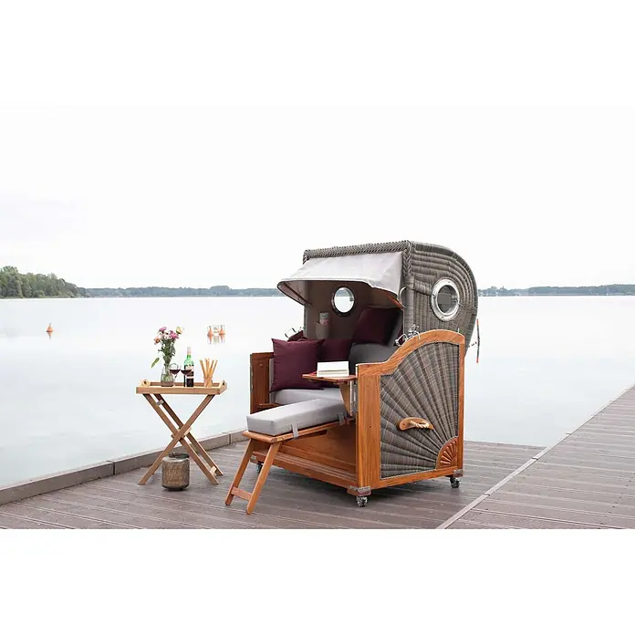 Bene Living Garten-Beistelltisch Norton - Teakholz, Klappbar, Rechteckig, Natur - Ein klassischer Strandkorb aus Korbgeflecht mit Holzelementen und grauen Polstern auf einem Steg am See, daneben ein Tisch mit Erfrischungen.
