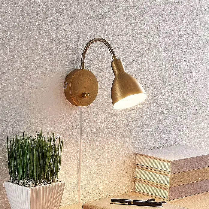 Lindby Wandleuchte Amrei - bronze / altmessing, bronze / altmessing, E14 - Messingfarbene Wandleuchte mit flexiblem Arm, an einer Wand montiert, beleuchtet einen Schreibtischbereich.