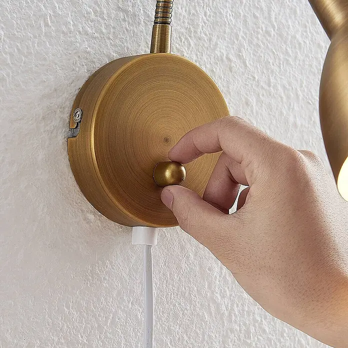 Lindby Wandleuchte Amrei - bronze / altmessing, bronze / altmessing, E14 - Hand dreht einen gebürsteten Messing-Drehdimmer an einer Wandleuchte mit weißem Stromkabel.