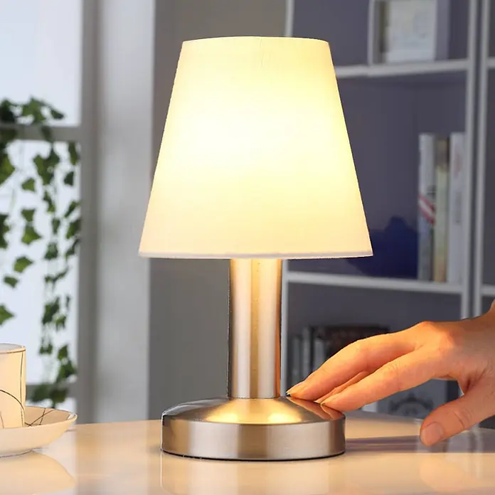 Lindby Tischleuchte Hanno - L x B x H: 14 x 14 x 24,5 cm, weiß / opal, E14 - Tischlampe aus gebürstetem Metall mit weißem Stoffschirm, eine Hand berührt den Fuß.