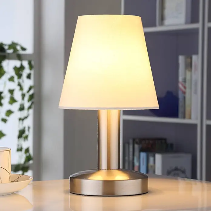 Lindby Tischleuchte Hanno - L x B x H: 14 x 14 x 24,5 cm, weiß / opal, E14 - Beleuchtete Tischlampe mit weißem Stoffschirm und gebürstetem Messingsockel, auf einem hellen Tisch stehend.