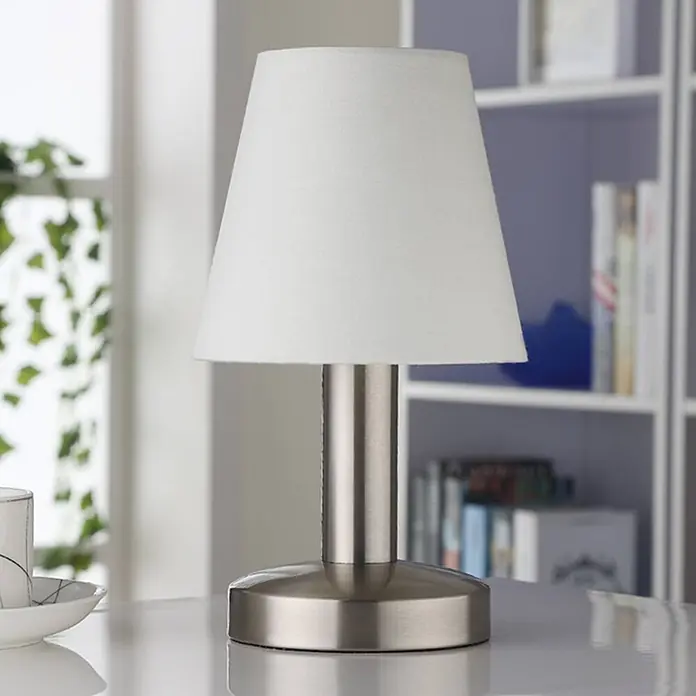 Lindby Tischleuchte Hanno - L x B x H: 14 x 14 x 24,5 cm, weiß / opal, E14 - Eine moderne Tischlampe mit weißem, konischem Stoffschirm und gebürstetem silbernem Metallsockel, auf einem Tisch neben einem Bücherregal.