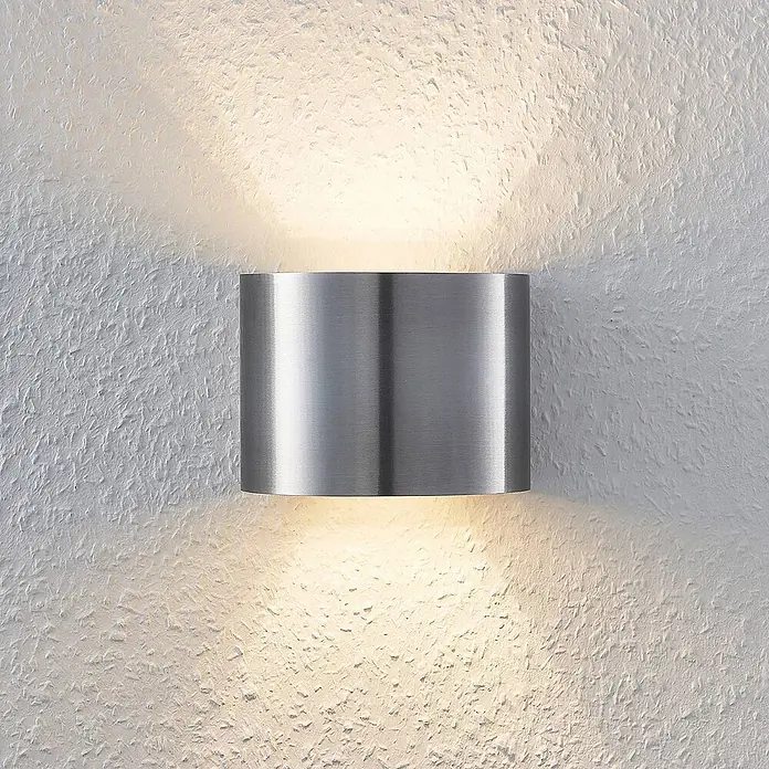 Arcchio Wandleuchte Zuzana - alu / grau / zink, G9 - Gebürstete Edelstahl-Zylinder-Wandleuchte, die warmes Licht nach oben und unten auf eine strukturierte Wand strahlt.