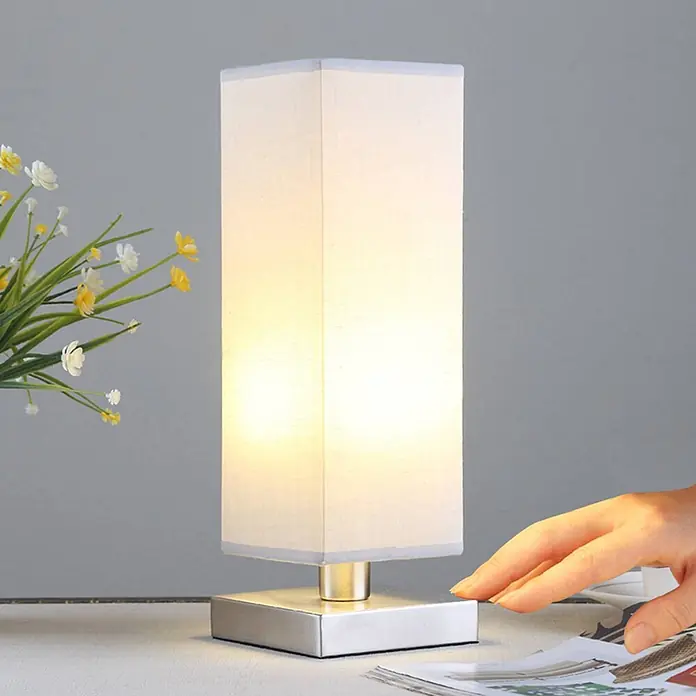 Lindby Tischleuchte Julina - L x B x H: 12 x 12 x 35 cm, alu / grau / zink, alu / grau / zink, E14 - Beleuchtete Tischleuchte mit rechteckigem weißen Stoffschirm und gebürstetem Metallsockel, eine Hand berührt den Sockel.