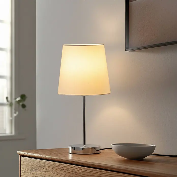 Lindby Tischleuchte Leza - chrom / silber, weiß / opal, E14 - Eine Tischlampe mit cremefarbenem Schirm und Chromfuß steht auf einer Holzkommode und spendet warmes Licht.